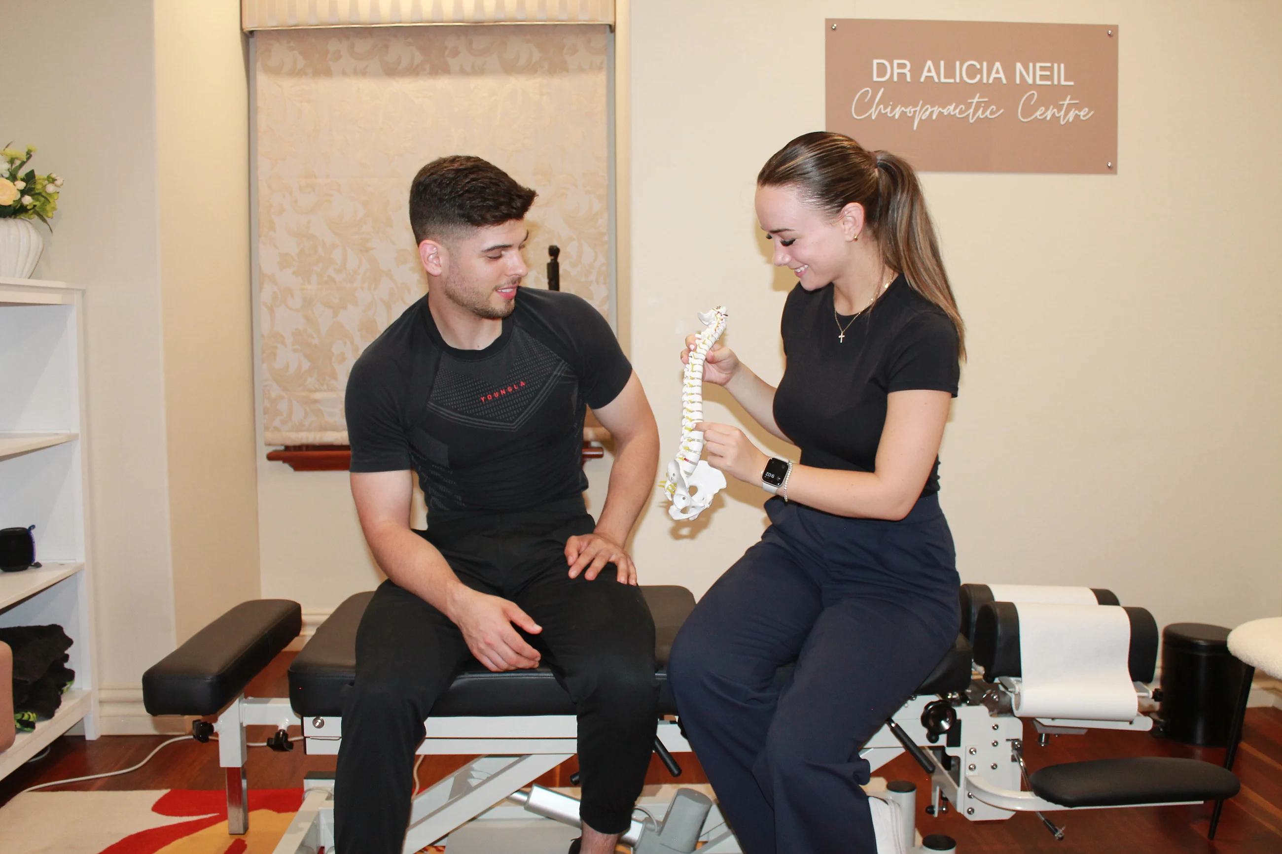 Alicia Neil smiling as she explains a model spine to a patient beside her Dr Alicia Neil Chiropractic Centre sign